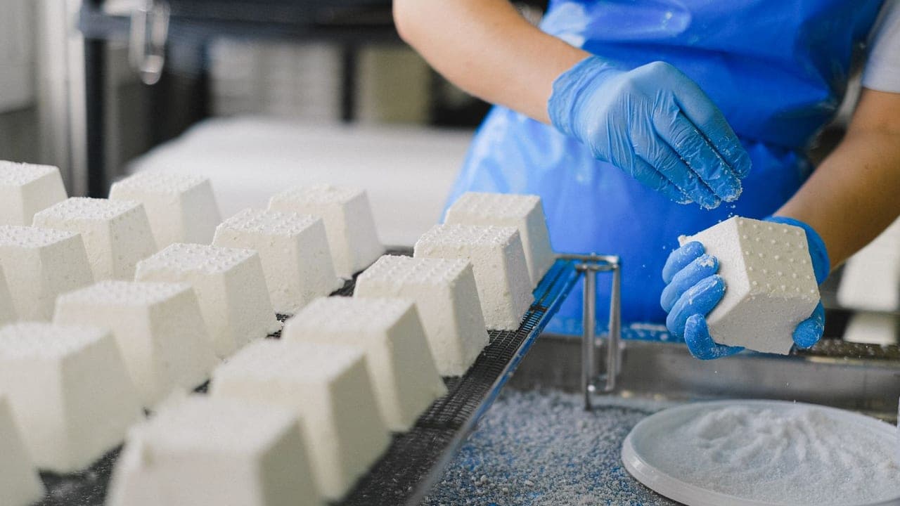 A dairy worker in blue gloves salting fresh cheese blocks on a draining rack